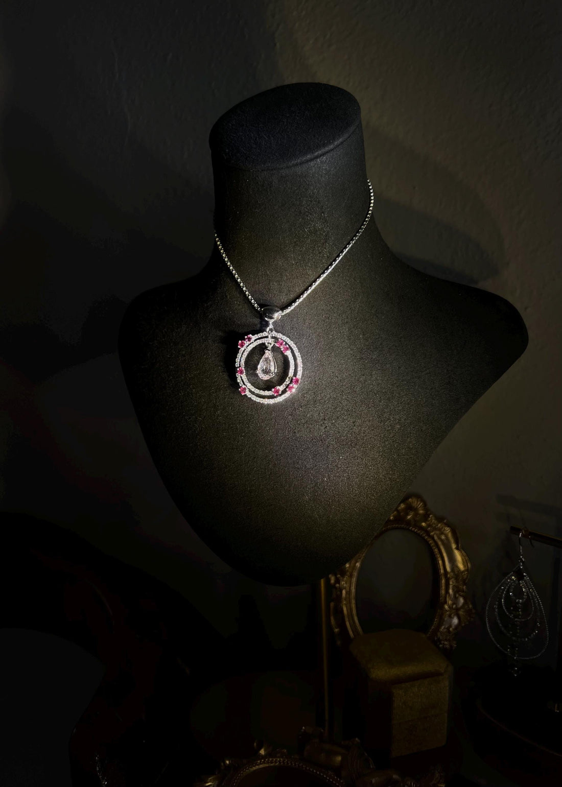 May include: Elegant pendant necklace displayed on a black jewelry stand. The necklace features a circular design with a pear-shaped gemstone in the center, surrounded by smaller pink gemstones and clear sparkling stones. The pendant hangs from a delicate chain. The overall style is sophisticated and luxurious.