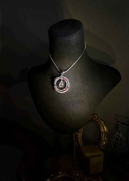 May include: Elegant pendant necklace displayed on a black jewelry stand. The necklace features a circular design with a pear-shaped gemstone in the center, surrounded by smaller pink gemstones and clear sparkling stones. The pendant hangs from a delicate chain. The overall style is sophisticated and luxurious.