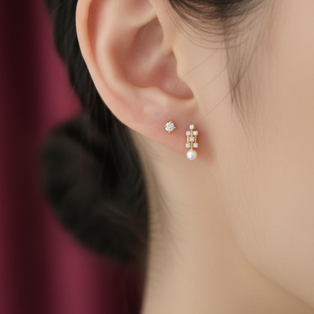May include: A close-up of a person's ear adorned with two earrings. One is a small, round stud with a central diamond surrounded by smaller stones. The other is a gold drop earring with a pearl and multiple small diamonds.