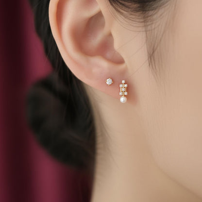 May include: A close-up of a person's ear adorned with two earrings. One is a small, round stud with a central diamond surrounded by smaller stones. The other is a gold drop earring with a pearl and multiple small diamonds.