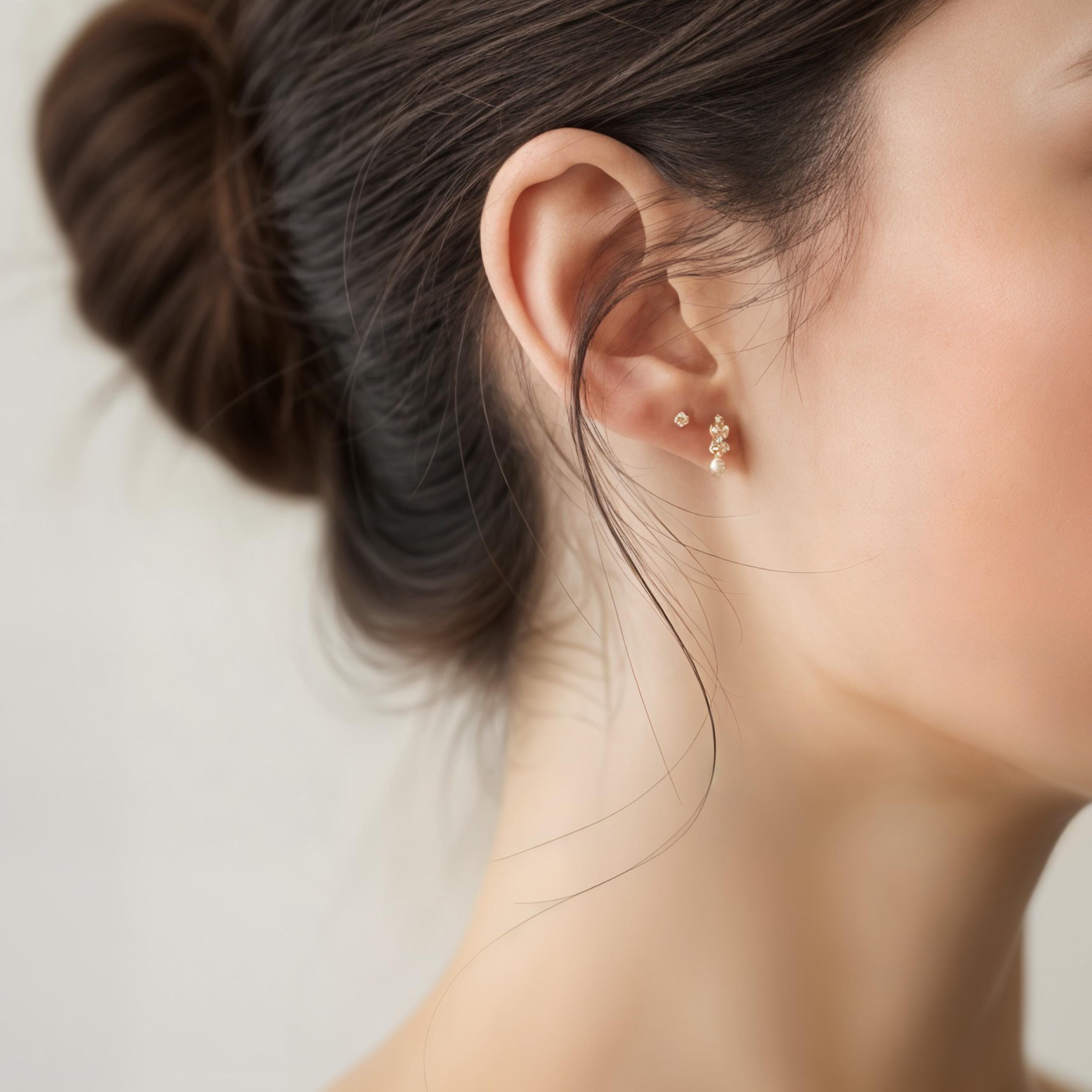 May include: Close-up of a woman's ear adorned with two delicate gold earrings. One is a small stud, and the other features a cluster of tiny pearls or crystals, creating a subtle, elegant sparkle. The woman's hair is styled in a bun.