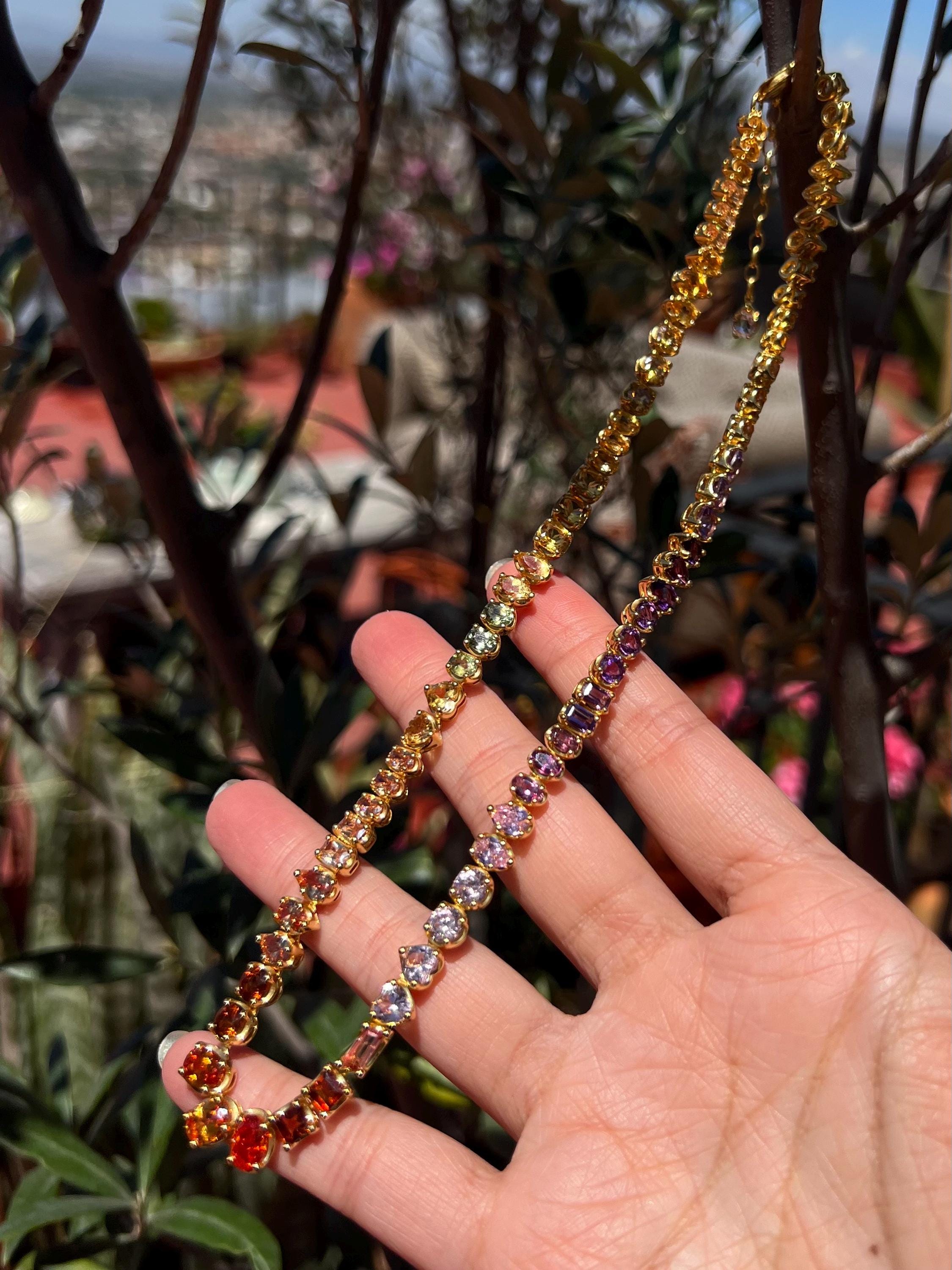 May include: A gold-toned necklace with a gradient of colorful gemstones. The gemstones transition from red to orange, yellow, green, pink, and purple. The necklace is held in a hand against a blurred background of greenery.